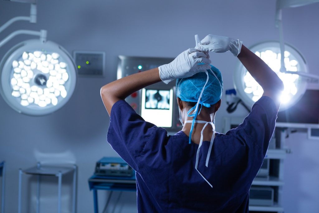 Rear view of young African-american female surgeon putting her surgical mask on in operating room at hospital. Shot in real medical hospital with doctors nurses and surgeons in authentic setting