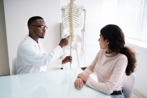 The image shows a Black doctor explaining aspects of the spine to a patient. The image visualizes when you should replace your cervical discs.