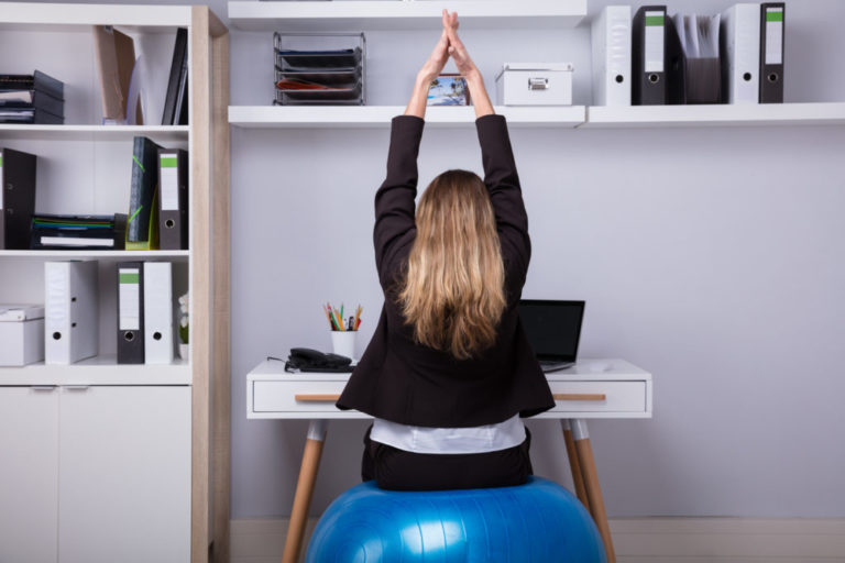 Rear View Of A Businesswoman Sitting On Fitness Ball Stretching Her Arms